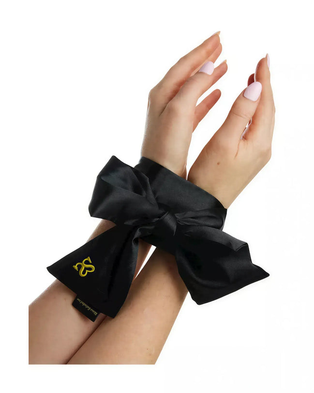 Black satin bondage ties with a large bow, secured around wrists, featuring a gold logo tag.